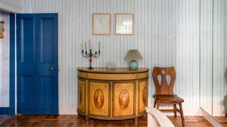 Bedroom with a blue painted door, striped blue wallpaper, a vintage console table with a painted design, an iron candelabra on top with colourful candles, and a vintage chair beside it