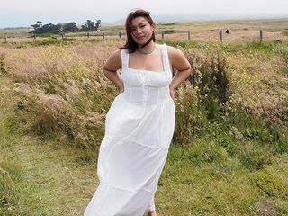 Marina wears white dress while standing in a field with the wind blowing.