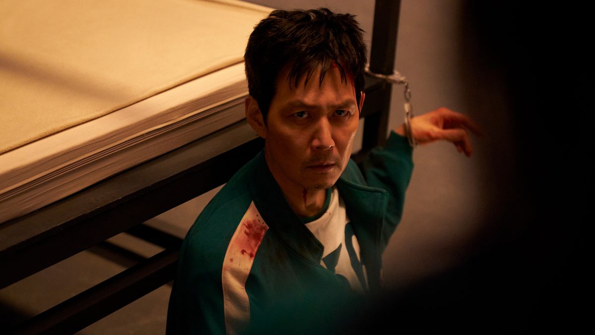 Squid Game season 3 player handcuffed to a bed with his green and white tracksuit covered in blood