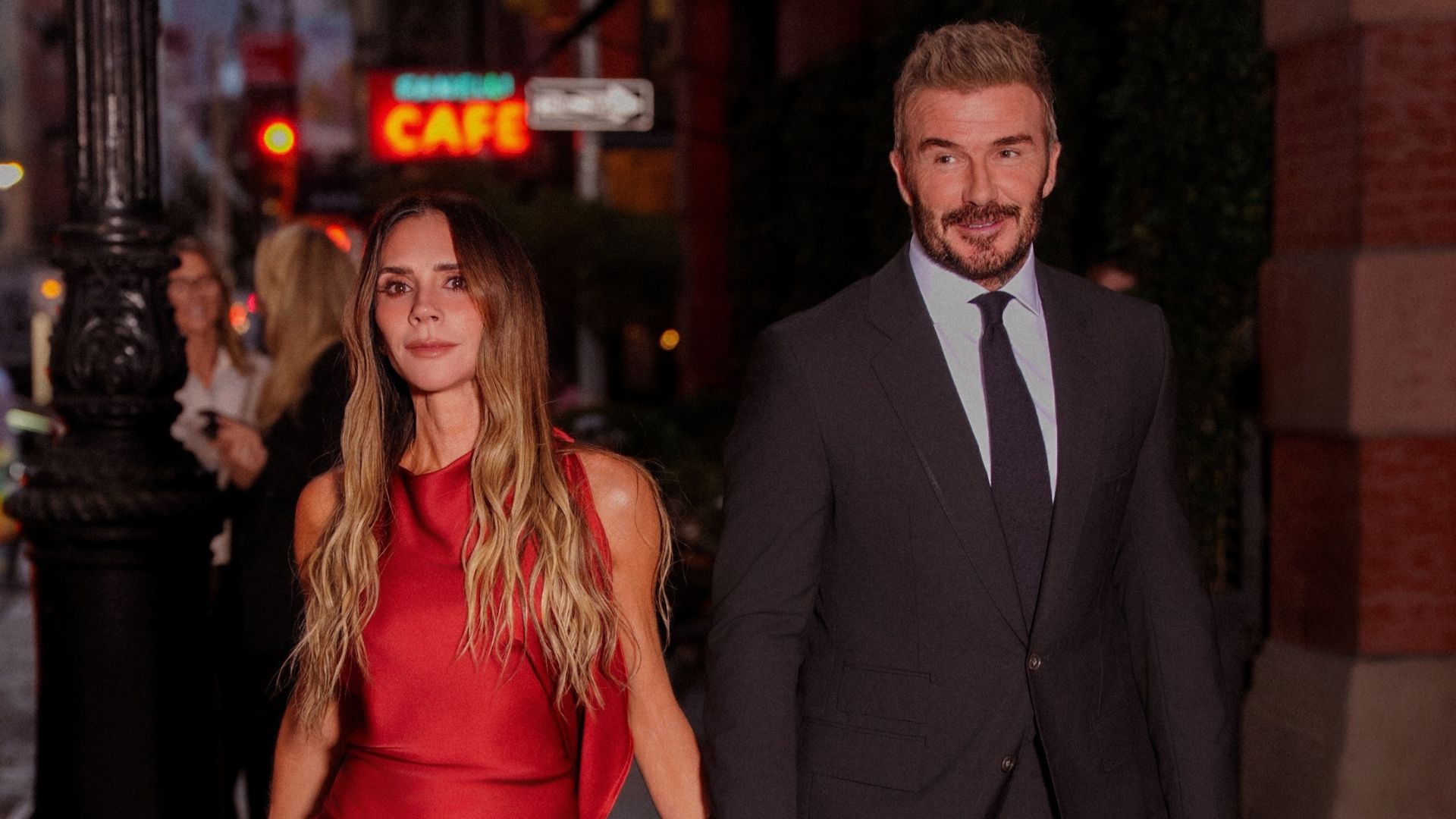 Victoria and David Beckham walk down the street holding hands. Victoria wears a red satin dress with plum heels, and David wears a suit.