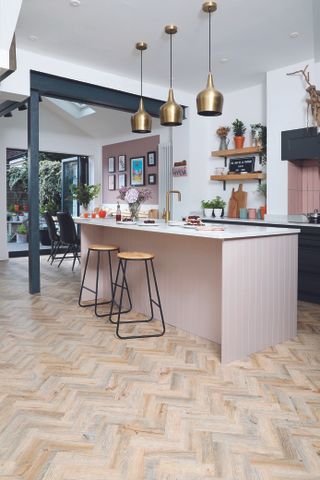 A light pink paneled kitchen island with white worktop, two wooden bar stools, and three large brass pendant lights above it