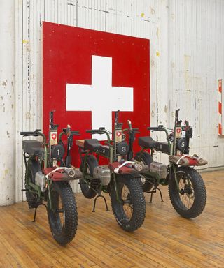 Installation view featuring three black electric motorcycles in a room with wooden flooring and distressed off-white coloured walls. There is a large piece of wall art behind the motorcycles of the red and white Swiss flag