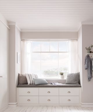 White reading nook positioned by window with wall paneling on ceiling, shaker-style drawers and assortment of white and grey cushions