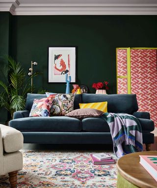 A dark green living area with a navy blue velvet couch with colorful throw pillows and throws, a floral patterned blue, pink, and cream rug, and a screen with red wavy panels and yellow bars