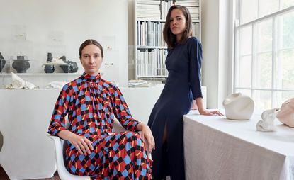 Artist Ekaterina Bazhenova-Yamasaki (sitting on a white chair and wearing a geometric dress) and jeweller Anna Jewsbury (Wearing a blue dress with thigh slits and resting on a white table beside Ekaterina) placed against a wide window in an artistic studio with scuptured around the room and a book shelf 