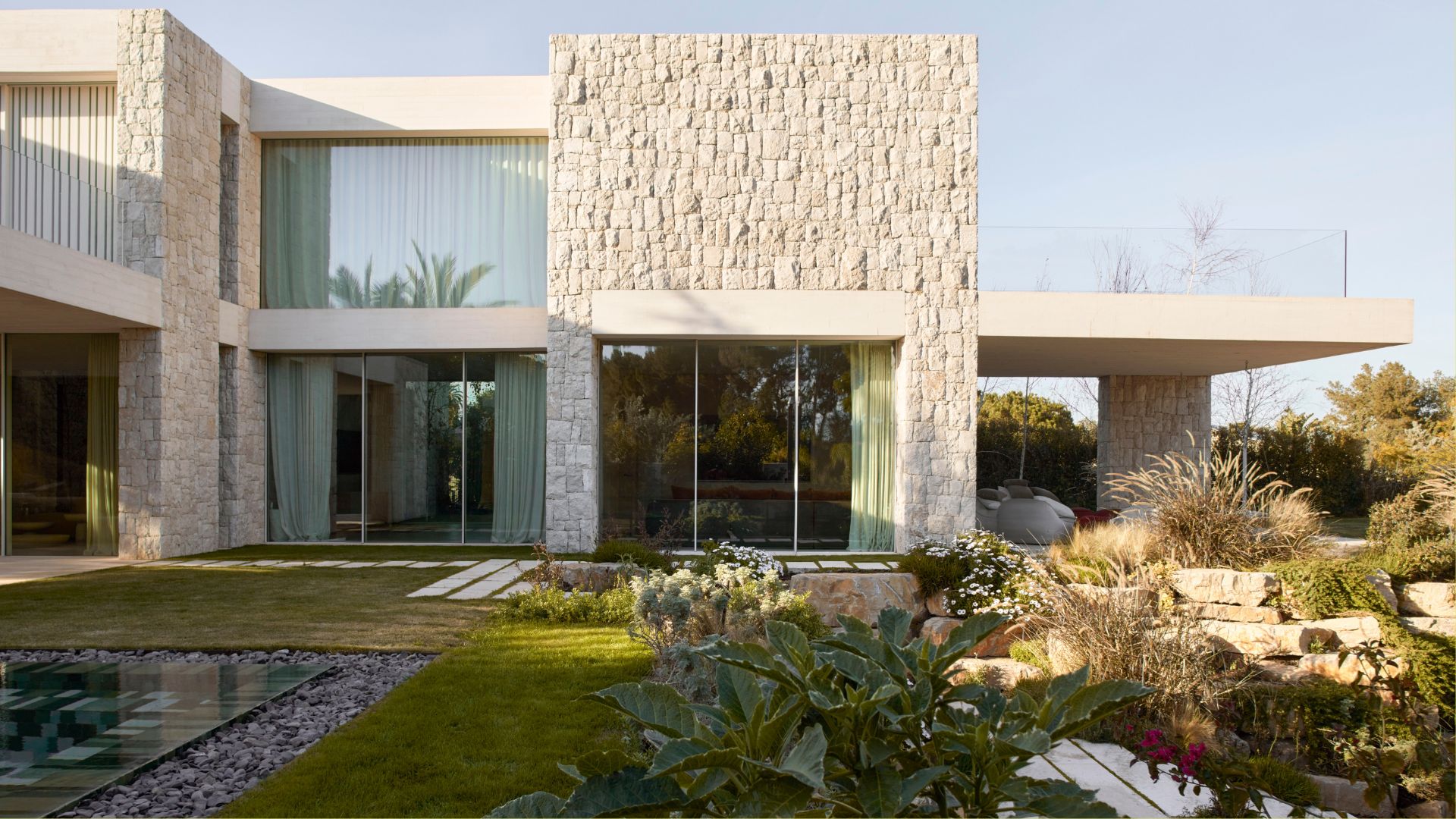 A backyard with a green lawn, a tiled pool, a textured facade, and lush landscaping
