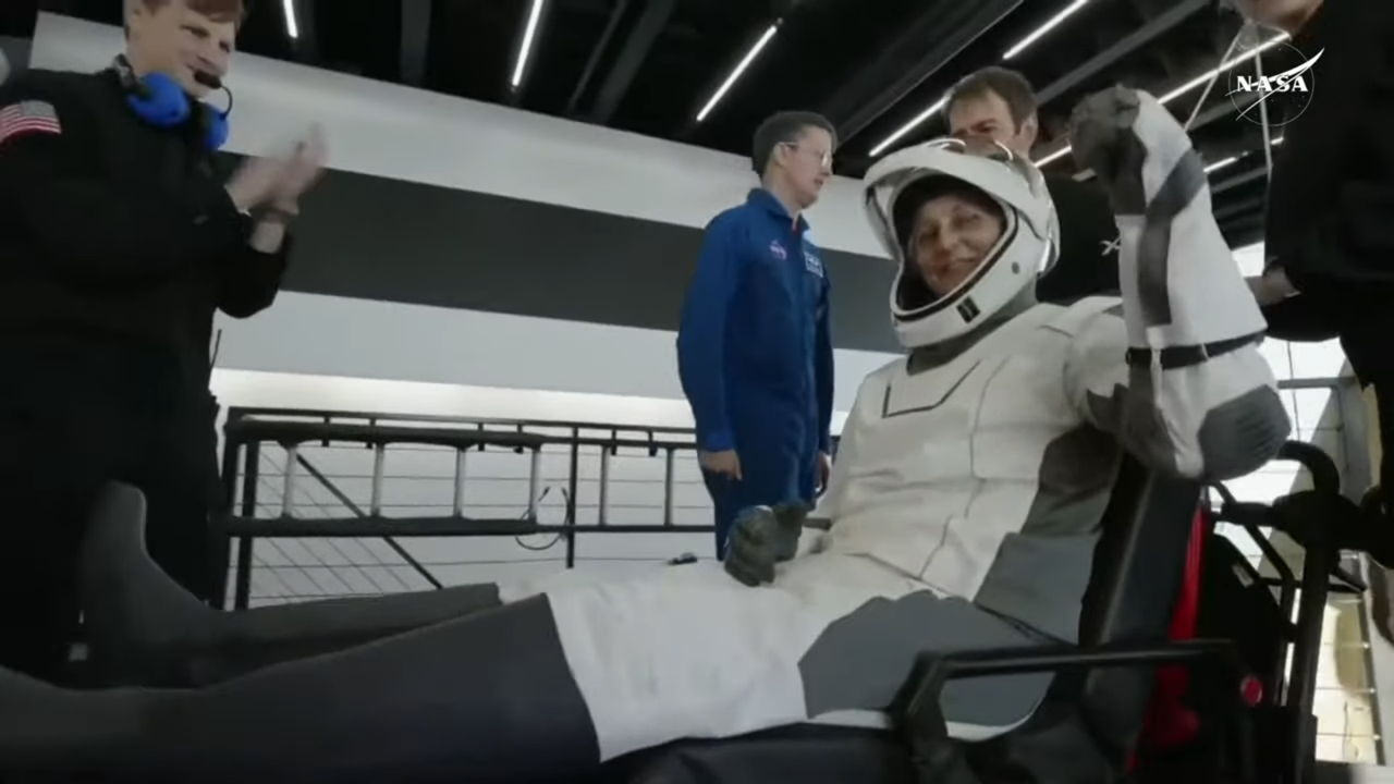 A female NASA astronaut in a white and black spacesuit waves after leaving their spacecraft on stretcher