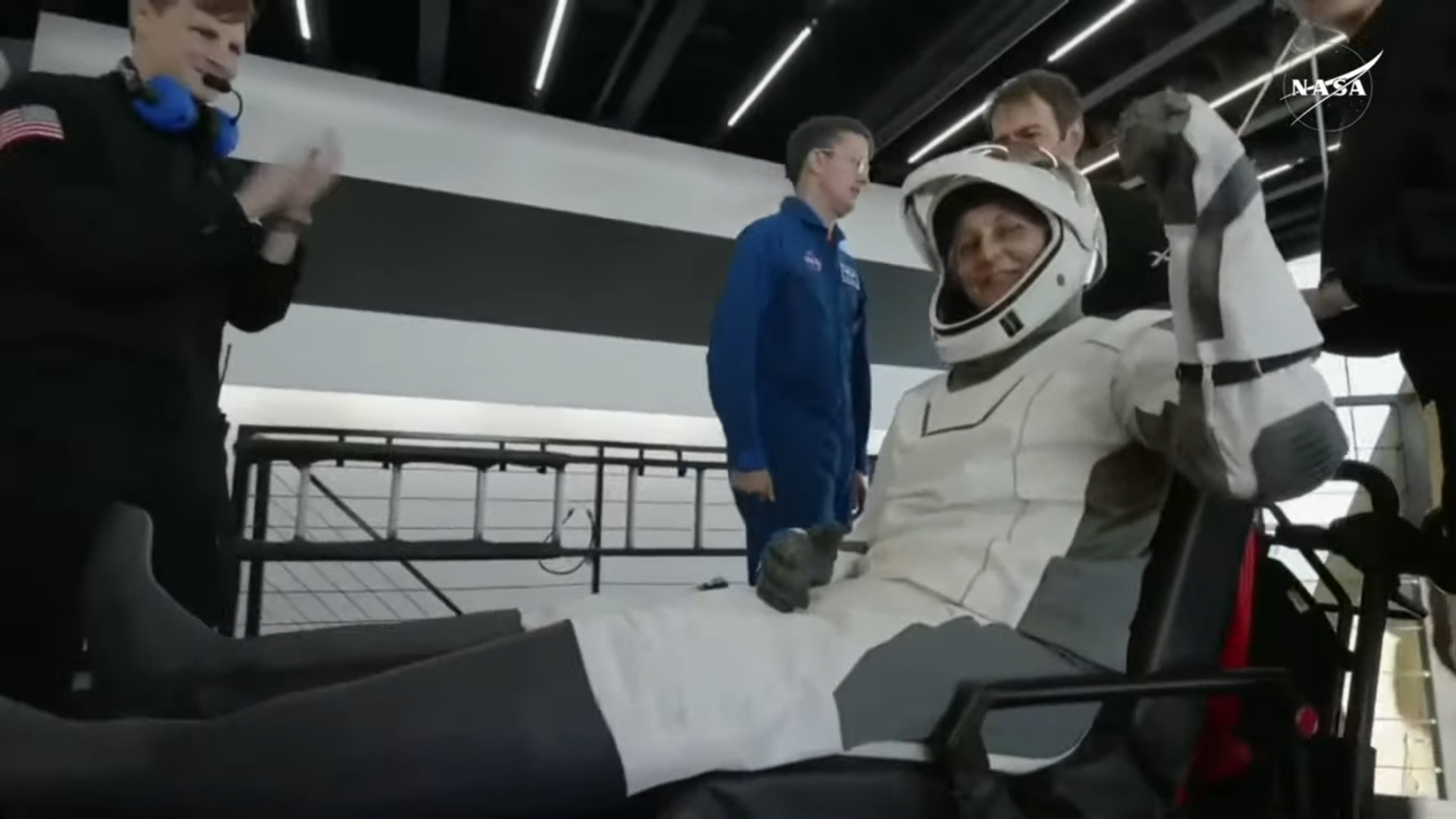 A female NASA astronaut in a white and black spacesuit waves after leaving their spacecraft on stretcher 