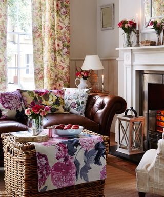 a neutral living room with bright floral curtains, cusions and throw, a wicker basket coffee table, with a brown leather sofa by a burning fireplace