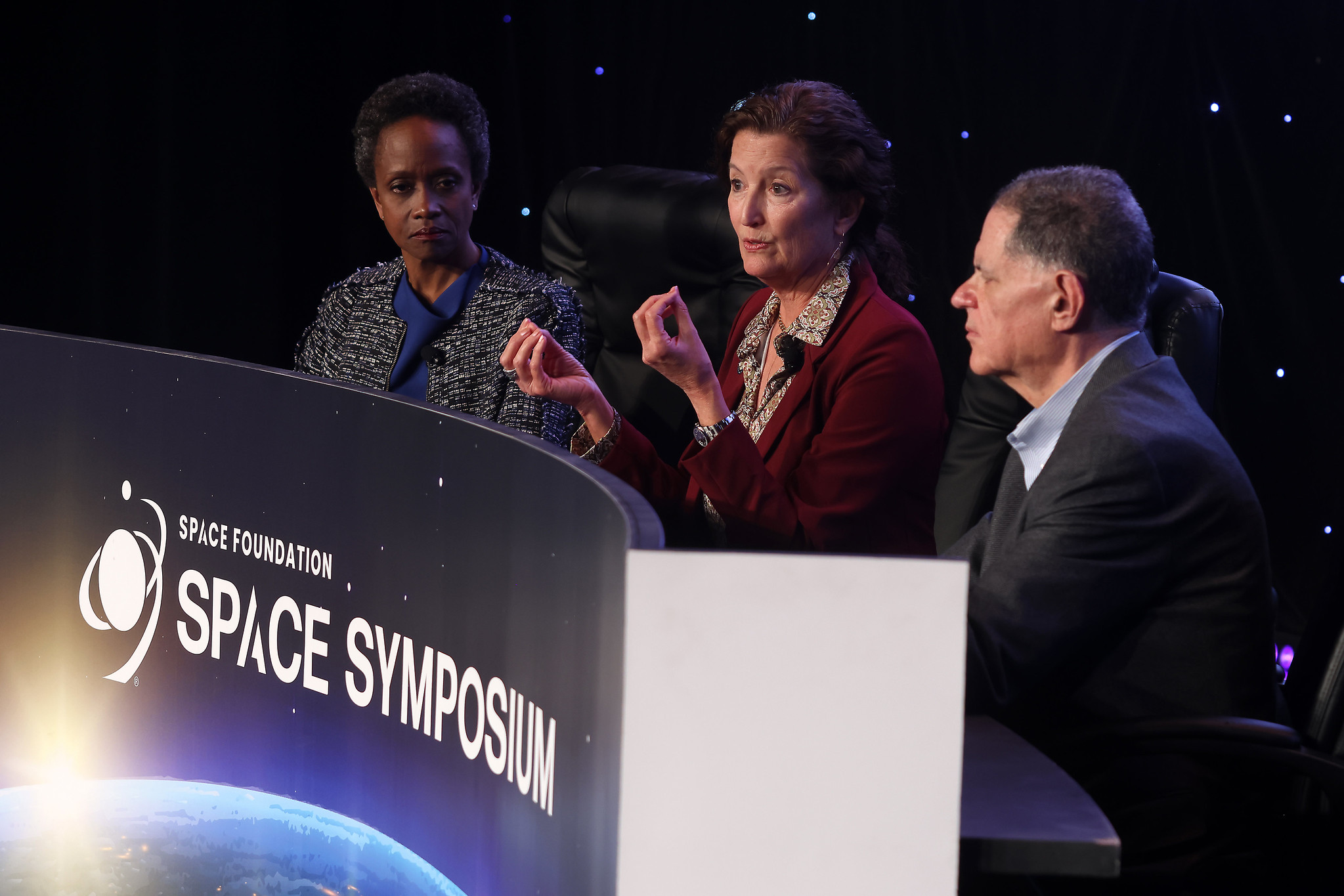 three people in business casual outfits sit at a table on a stage in front of a backdrop that resembles the stars
