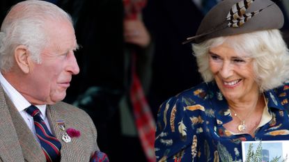 King Charles wearing a tweed jacket and vest smiling at Queen Camilla wearing a blue feather print dress and holding a sprig of greens