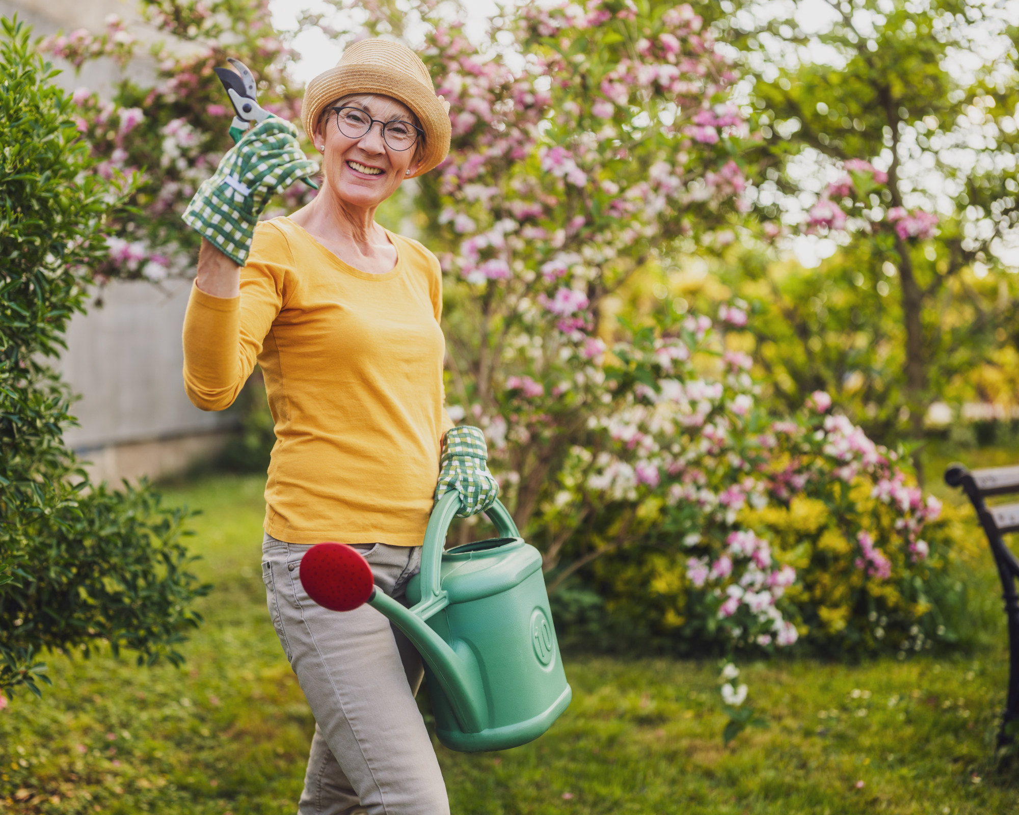 Woman wearing a yellow top and green gardening gloves in a garden, with hand pruners and a watering can