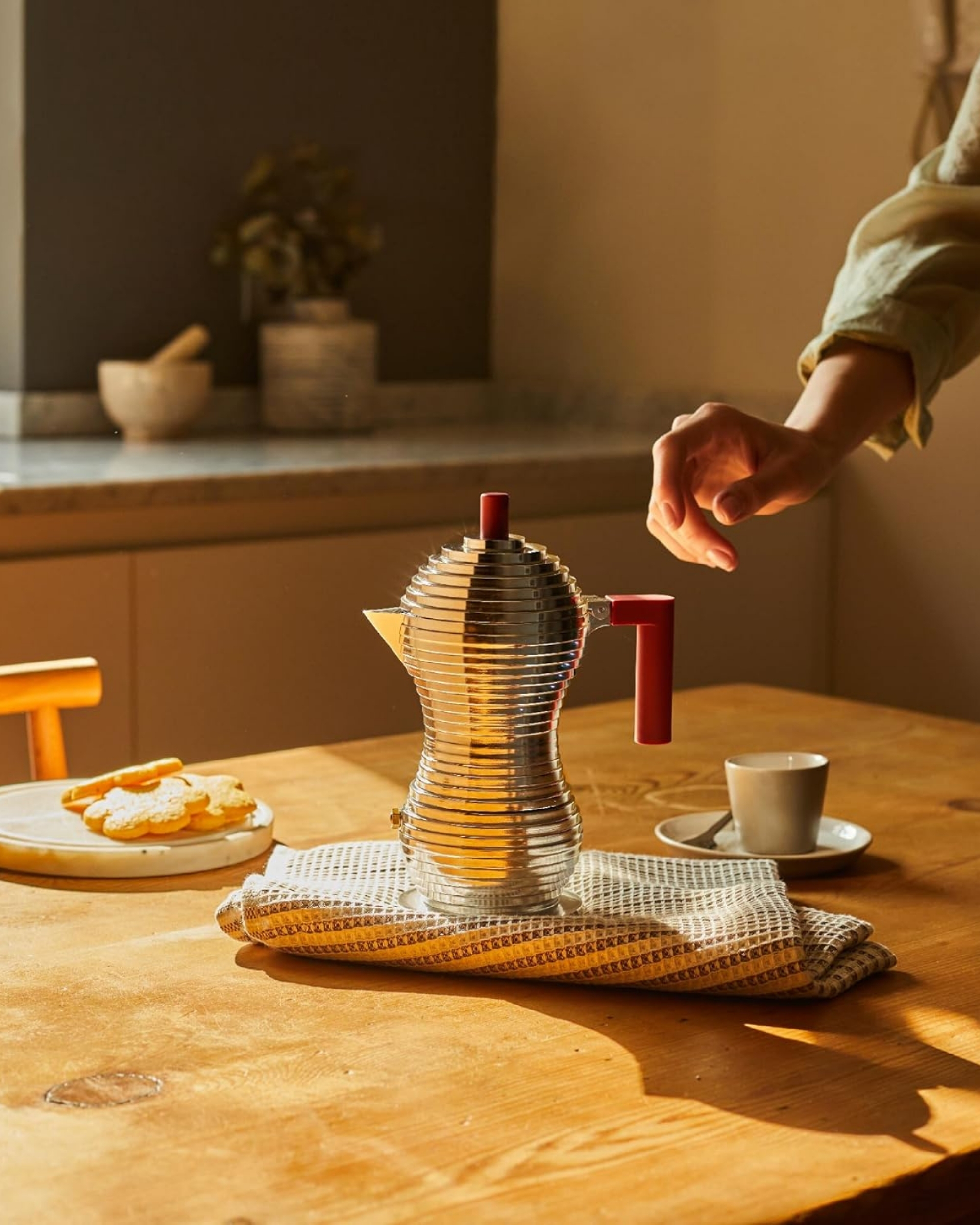 Image of a silver and red, modern moka pot sitting on a towel on a wooden table. There is a hand reaching for the moka pot.