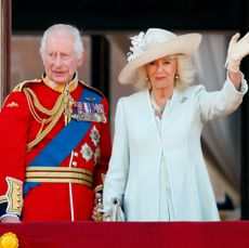 King Charles in a red uniform and Queen Camilla in a blue coat dress standing on the balcony of Buckingham Palace 