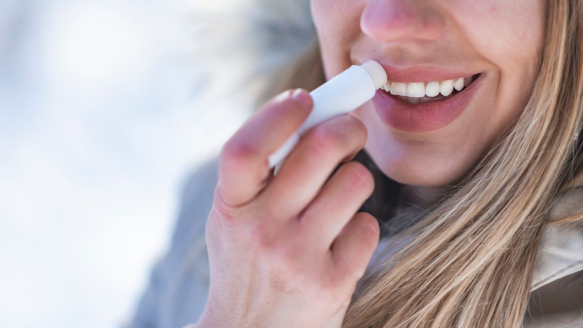 close up photo of a smiling woman standing outside in a coat and applying lip balm 