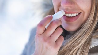 close up photo of a smiling woman standing outside in a coat and applying lip balm 