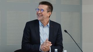 Professor Virginijus Šikšnys smiles in a blue suit as he's seated in front of a microphone during a talk
