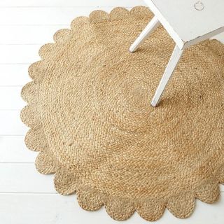 A circular woven jute rug with a scalloped edge displayed on the floor under a white dining table chair. 