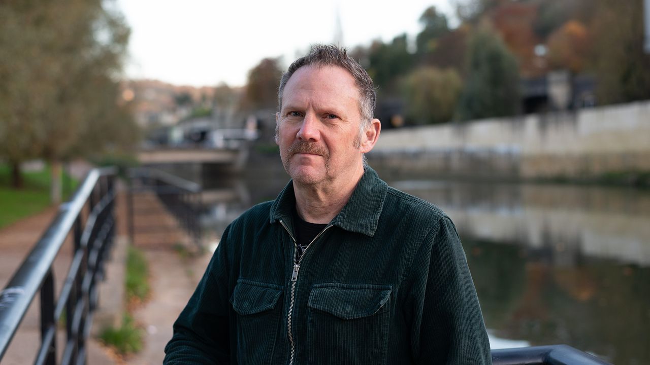 Man with green jacket and handlebar moustache, stood in front of river