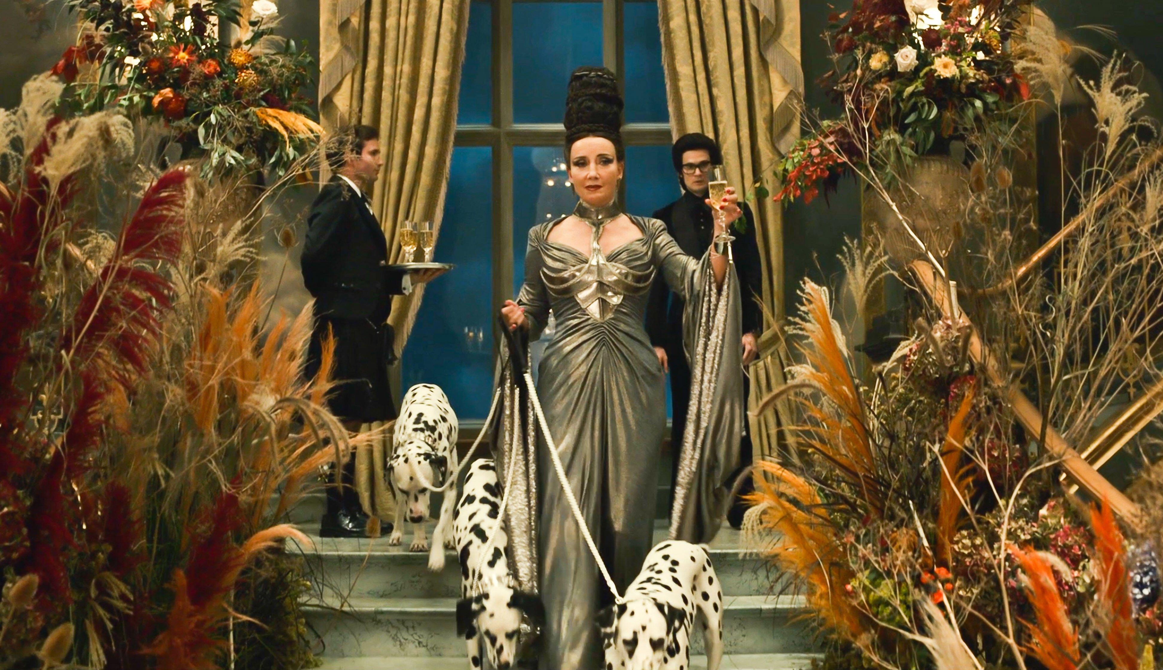 Curella film A glamorous woman in a silver evening gown descends a grand staircase holding champagne, flanked by two dalmatians on leads amid lavish floral arrangements