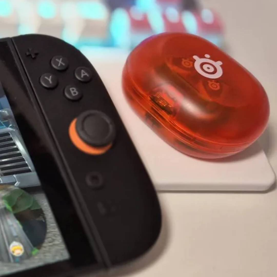Orange SteelSeries Arctis Game Buds next to Switch 2 on white desk.