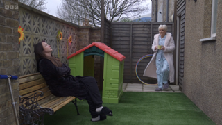 Stacey Slater wearing a dressing gown sitting on a bench in her garden with a children's playhouse. Jean Slater walks over with a cup of tea.