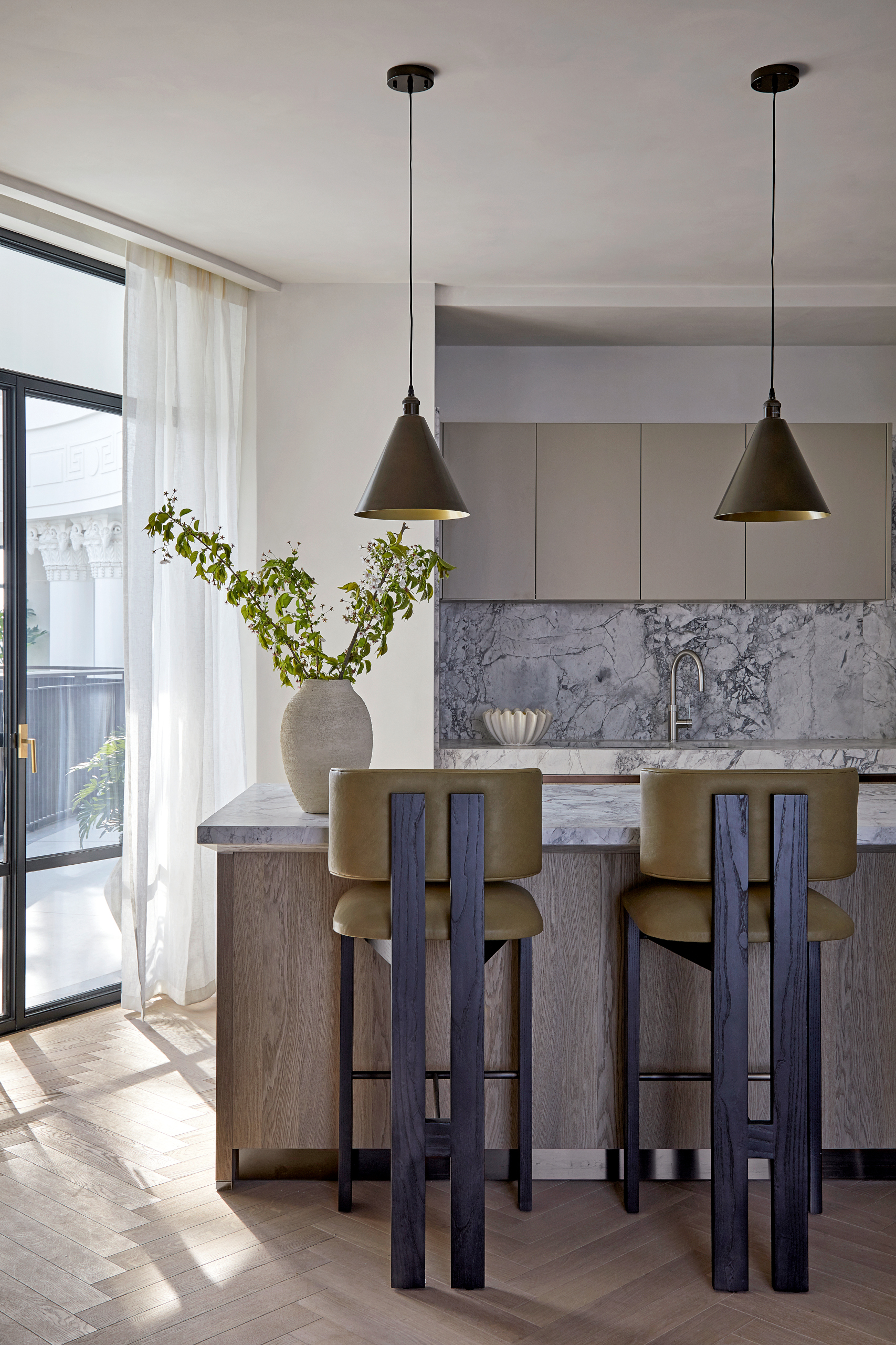 Kitchen with blue-veined marble backsplash, wood units and island, black wood bar stools with olive seat pads and dark bronze pendant lights