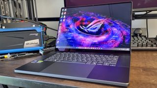 An Asus ROG Zephyrus G16 (2026) gaming laptop on a wooden-topped table next to a benchmarking rig