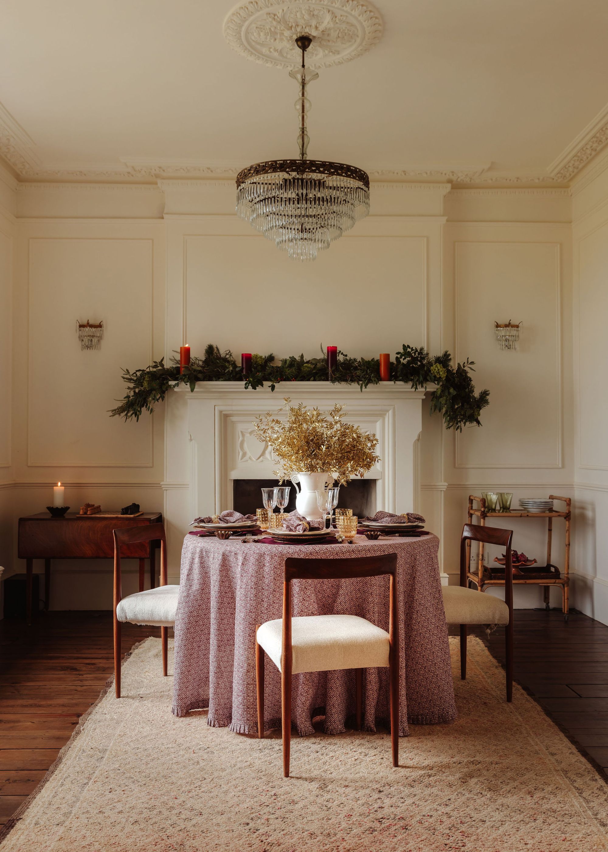 A dining room with a round dining table with a muted pink tablecloth. A festive garland on the fireplace and candles throughout the room.