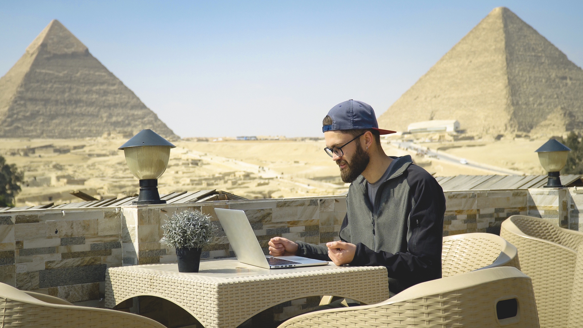Young man works from caf&eacute; near Egyptian Pyramids, typing into a laptop computer from and outdoor table.