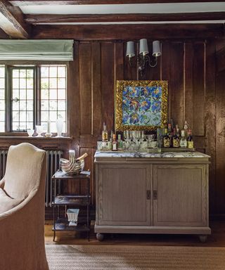 A room with a carved wood panelled wall and a covered dining chair, a sideboard and drinks tray, and an artwork in a gilded frame. Exposed beams and rug on the floor.