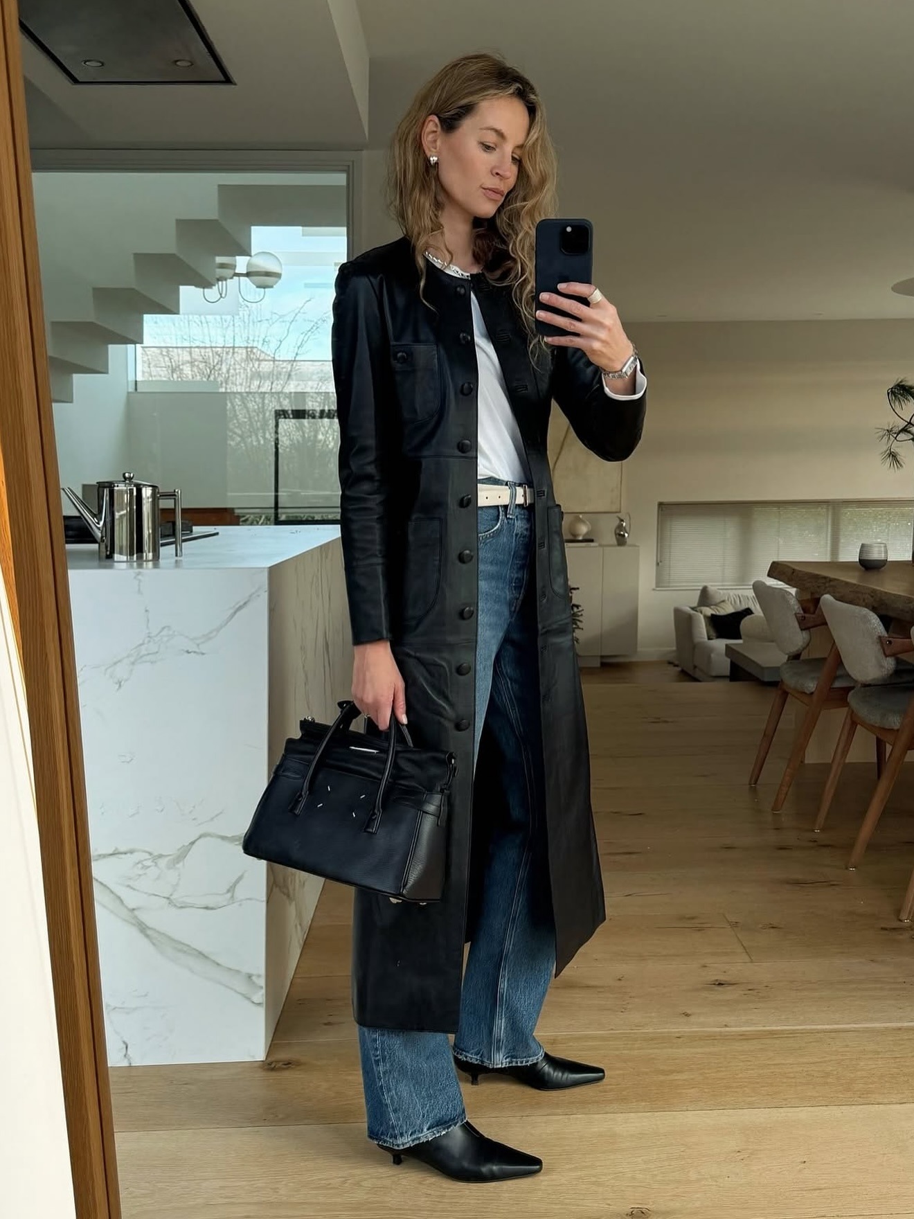 Influencer wears a collarless black leather trench, a white tee, blue jeans, and kitten-heel boots.