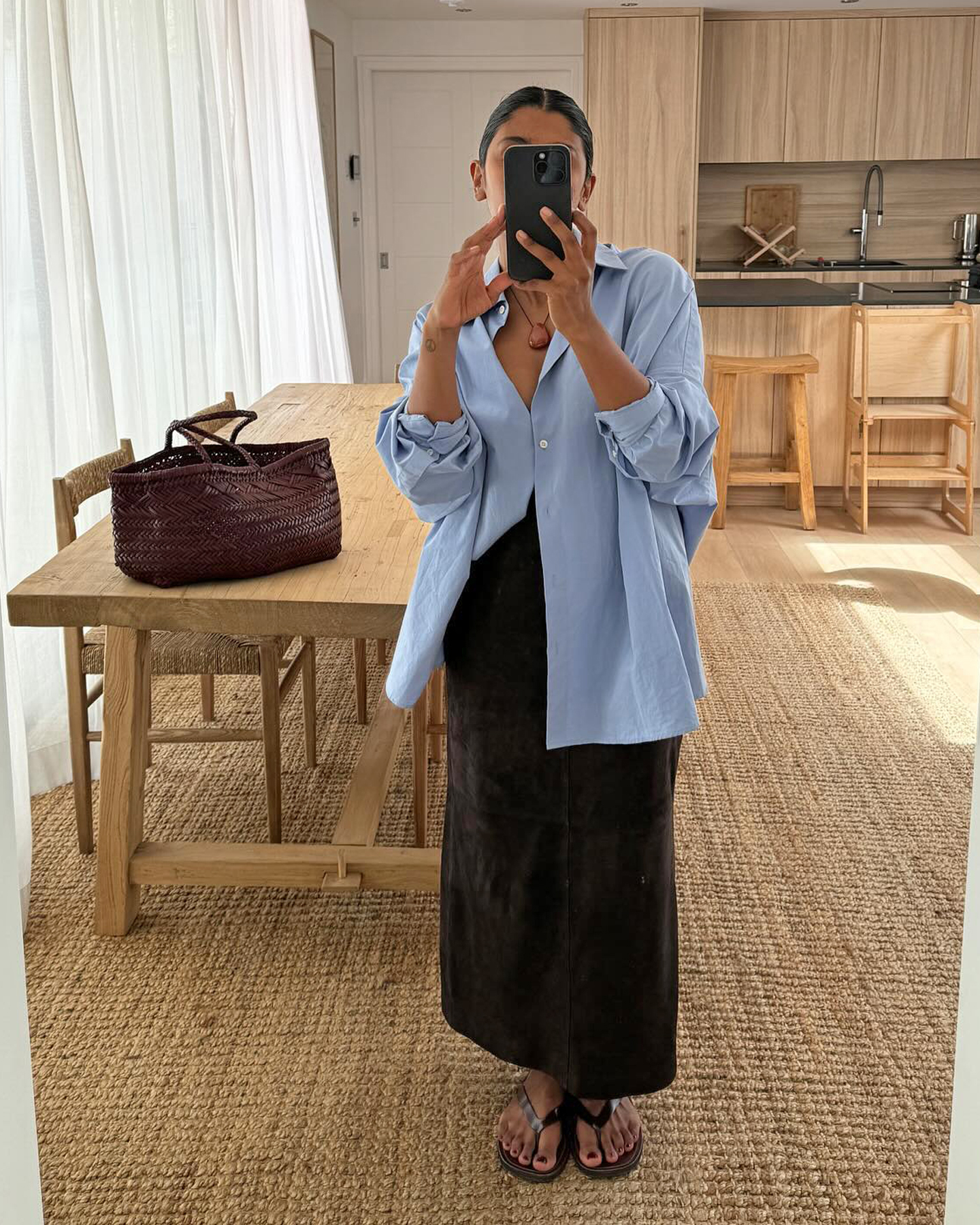 British style influencer Monikh poses for a mirror selfie in her chic London home wearing an oversize blue button-down shirt, black maxi skirt, black flip-flop sandals, and a brown woven leather tote bag