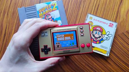 Hand holding Mario Game and Watch handheld with Super Mario 2 NES cartridge and Japanese Disc System game on woodgrain table.