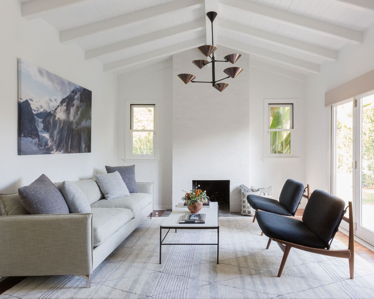 Light and bright living room painted white, wooden ceiling painted white, dark wood flooring covered in a large cream and light gray patterned rug, large light gray sofa, two matching wooden lounge chairs with dark gray upholstery, coffee table with black frame and marble tabletop, brick fireplace painted white, artwork above sofa, metallic pendant light