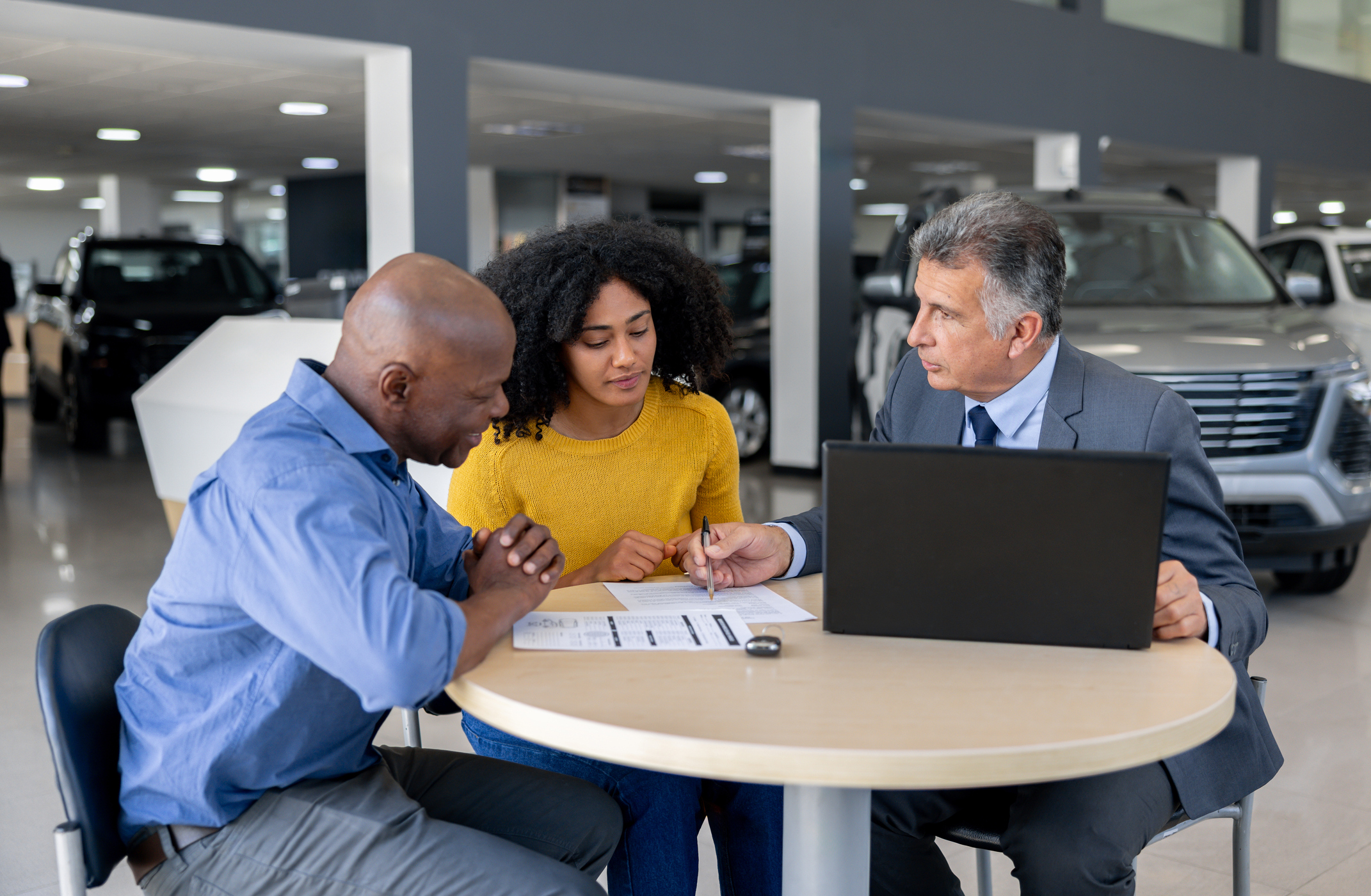 A car salesman discusses a contract with potential buyers while seated at a table inside a dealership.