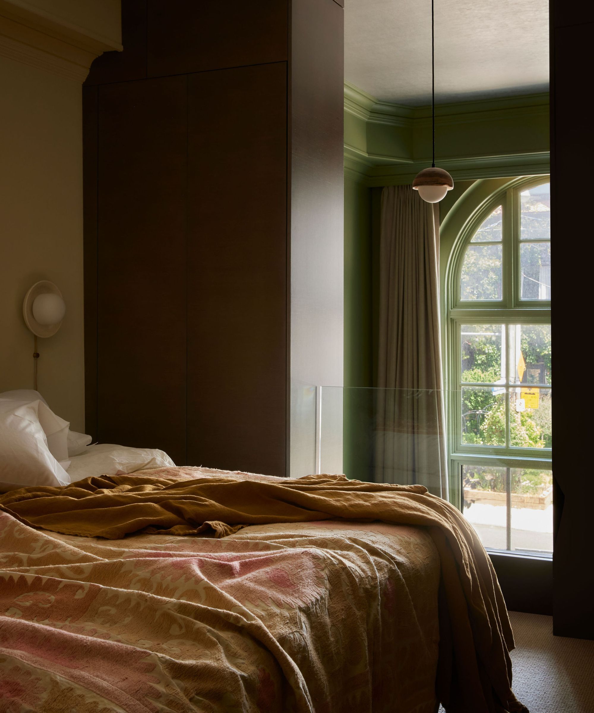 A bedroom with a gold and pink patterned bedspread, dark brown built-in storage, and a large arched window with green trim