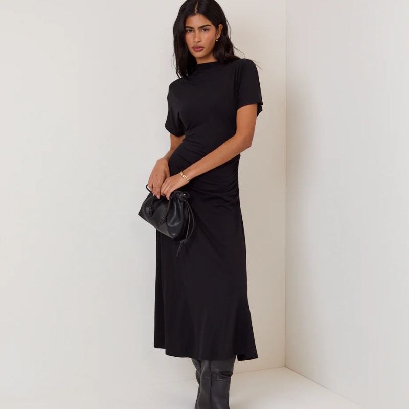 A woman stands in a corner wearing a black Nobody's Child dress and holding a clutch