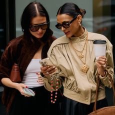 street style photo from New York Fashion Week of two women in chic fall outfits looking at a phone with a strawberry phone chain