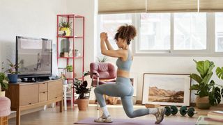 Woman learning how to do squats in her living room; she is working on a yoga mat and behind her sits a pair of dumbbells