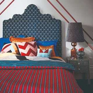 A bedroom with a bed with a large, curved printed headboard, a red striped bedspread and a leopard-print lamp