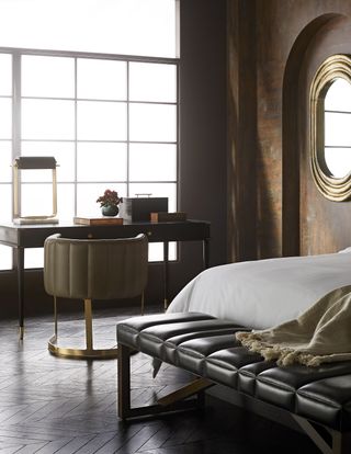 bedroom home office with black desk at window, herringbone floor, leather footstool, bed on the right, large oval gold mirror