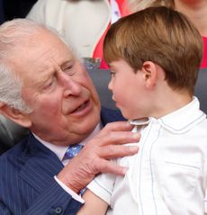 King Charles sitting in a chair wearing a blue suit and a blue tie with Prince Louis, dressed in a white shirt, on his lap 