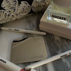 A flatlay picture of the Caia x Rotate makeup collaboration with a handbag and makeup products and a perfume bottle