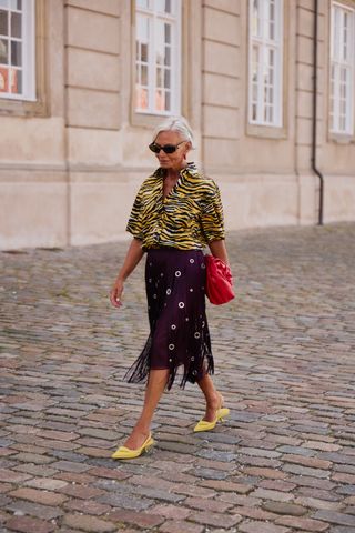 A woman wearing black sunglasses, a yellow zebra-print polo shirt, a burgundy fringe skirt with silver grommets, yellow prada slingback heels, and a large red clutch.