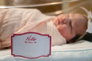 A newborn baby in a cot with a label reading 'Hello, my name is'