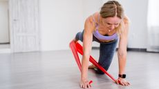 Woman exercising in empty room. She is in on all fours with one leg raised behind her, with a resistance band held under each hand and looped around her lifted foot.