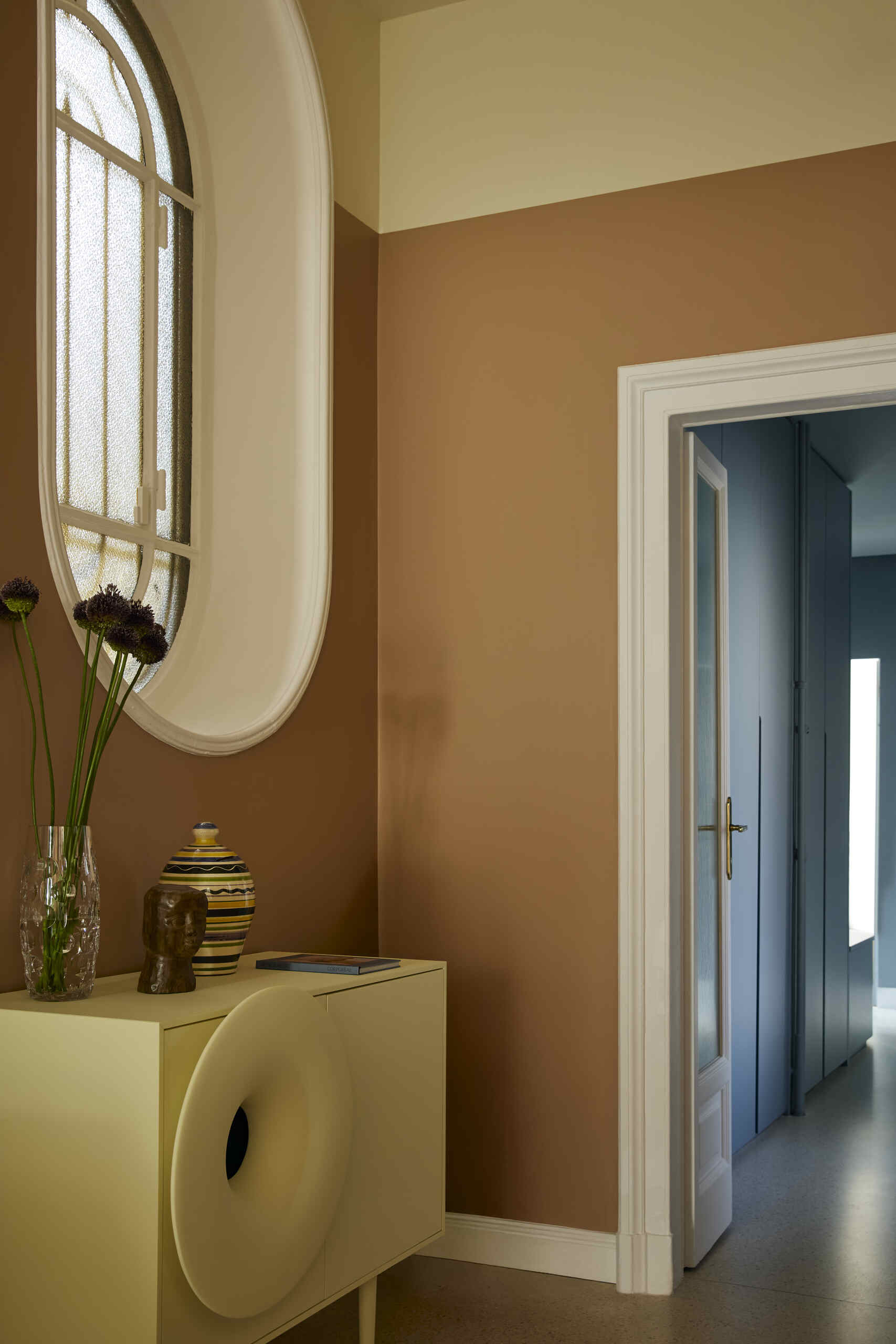 A burnt orange entryway with an oval-shaped window and a light yellow cabinet under the window. There is a door that leads into a light blue hallway.