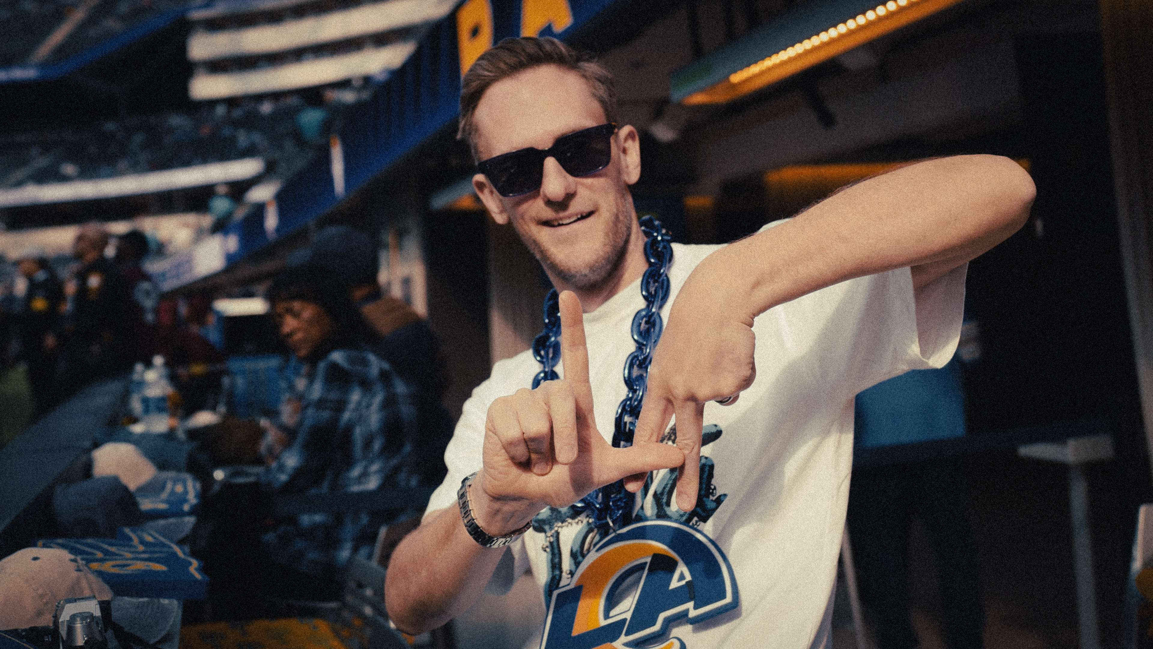 Harry Ead wears sunglasses and a Rams t-shirt while making a hand gesture at a sports stadium.