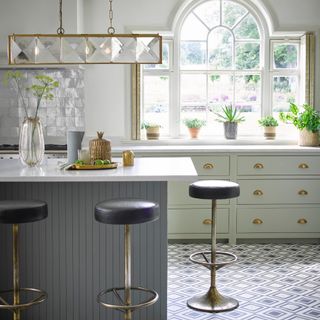 white and pale green kitchen with grey kitchen island, patterned floor tiles, black and brass bar stools, brass and glass pendant over island, brass handles, plants